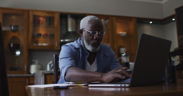 Senior man working on a laptop in home kitchen with glasses, focused on his task. This could be used for themes such as remote work, technology use among older adults, or home office environments. Suitable for websites and articles regarding working from home, productive routines, and modern senior lifestyle.