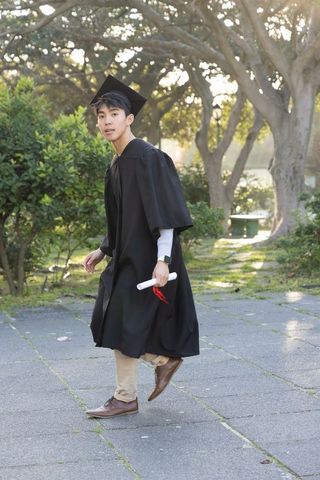 Young asian graduate walking on campus in cap and gown holding diploma