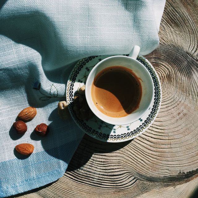 Sunlight casts a warm glow on cup of espresso placed on rustic wooden table. Various nuts like almonds are spread nearby. Ideal for marketing coffee products, cafes, lifestyle blogs, morning routines, or relaxation themes.