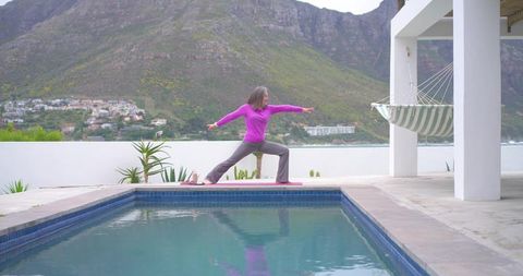 Woman practicing warrior ii on poolside terrace with mountain and ocean view