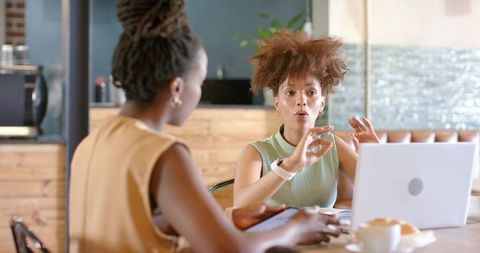 Two black women collaborating in cafe, gesturing over laptop and tablet during meeting