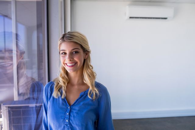 Young female executive standing near a window in a modern office, smiling confidently. Ideal for business, corporate, and professional themes. Can be used in articles, presentations, and websites related to workplace environments, career success, and professional development.