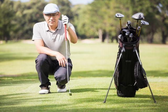 Senior golfer crouching on a golf course, posing with golf club and bag of clubs. Ideal for use in articles or advertisements related to golfing, senior activities, outdoor sports, and healthy lifestyles.