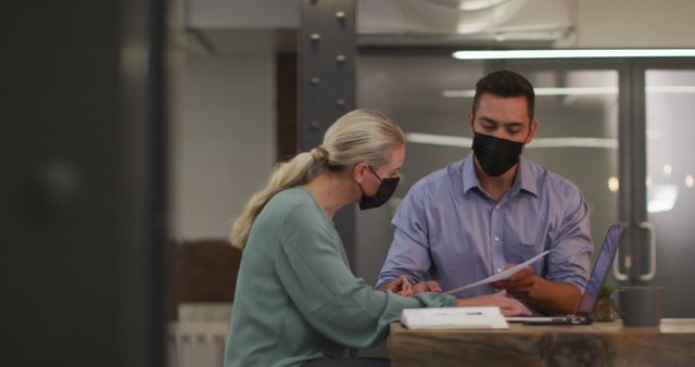Two coworkers sitting at a desk in a modern office, wearing masks and collaborating on a project. They are both looking at documents and laptop, immersed in discussion. Suitable for themes around business teamwork, pandemic impact on work, office safety, remote and hybrid work strategies.