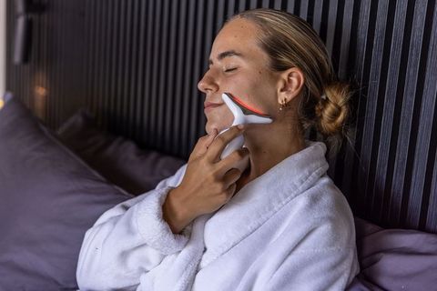 Woman using red light therapy facial massager while relaxing in white bathrobe on bed