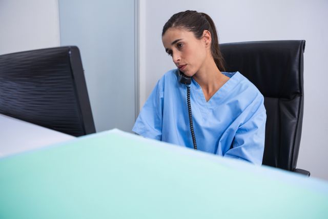 Doctor in blue scrubs talking on phone while looking at computer at desk in clinic. Ideal for use in healthcare, medical, and professional communication contexts. Suitable for illustrating medical consultations, healthcare technology, and office work in a clinical setting.