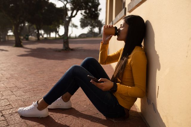 Front view of a biracial woman enjoying free time in nature on a sunny day, sitting on the ground, leaning against the wall, drinking coffee and holding a smartphone.