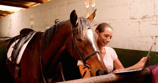A young Caucasian woman interacts with a horse while holding a laptop, sharing a moment of relaxation or work with the animal. Her smile suggests a bond with the horse, highlighting the connection between humans and animals.