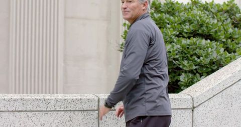 Senior man checking watch while standing by urban granite balustrade with greenery
