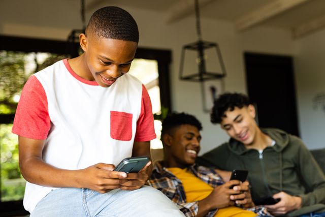 Three diverse teenage boys are sitting together at home, each engaged with their smartphones. They are smiling and enjoying their time, indicating a relaxed and friendly atmosphere. This image can be used to illustrate themes of modern technology, social media interaction, teenage lifestyle, and friendship. It is suitable for articles, advertisements, and social media posts related to youth culture, technology, or social bonding.