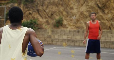 Holding blue basketball facing defender on outdoor streetball court near hillside