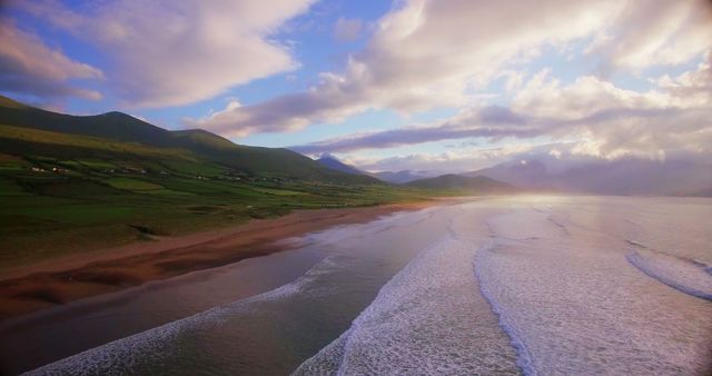 A serene coastal landscape unfolds with a vast beachfront and rolling hills under a dramatic sky at sunset. The interplay of light and shadow over the natural scenery creates a peaceful and picturesque setting.