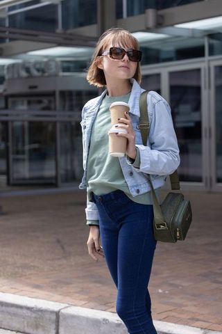Young woman with takeaway coffee and sunglasses near modern glass building, urban commuter