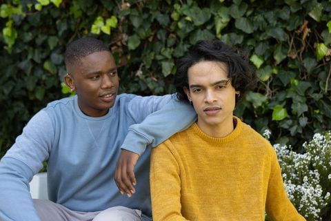 Black and mixed-race young men relaxing on garden bench wearing cozy sweaters, candid friendship