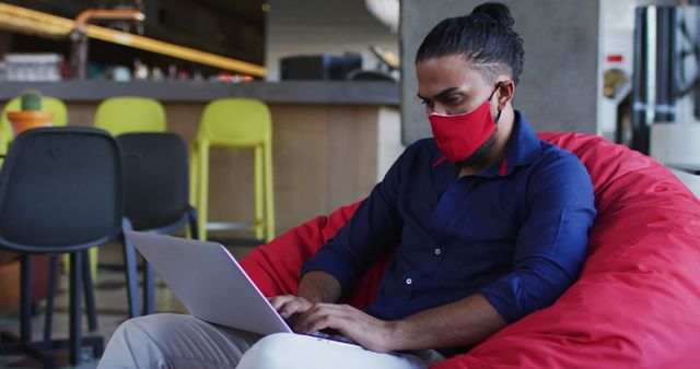 Man wearing face mask working on laptop in modern cafe. Useful for illustrating remote work, pandemic safety measures, digital nomad lifestyle, and business productivity. Ideal for blogs, articles, and promotions related to technology, working from home, or contemporary work environments.