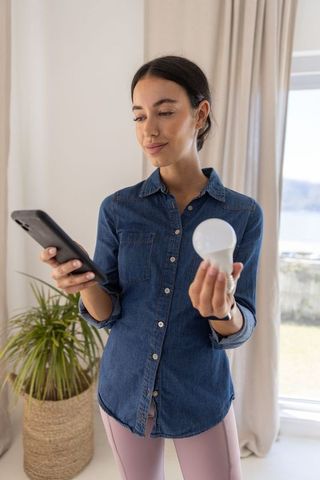 Woman holding smartphone inspecting led bulb in bright minimal home interior