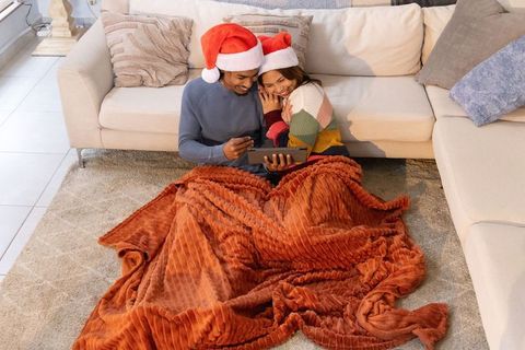 Cozy diverse couple wearing santa hats snuggling under rust blanket while browsing tablet