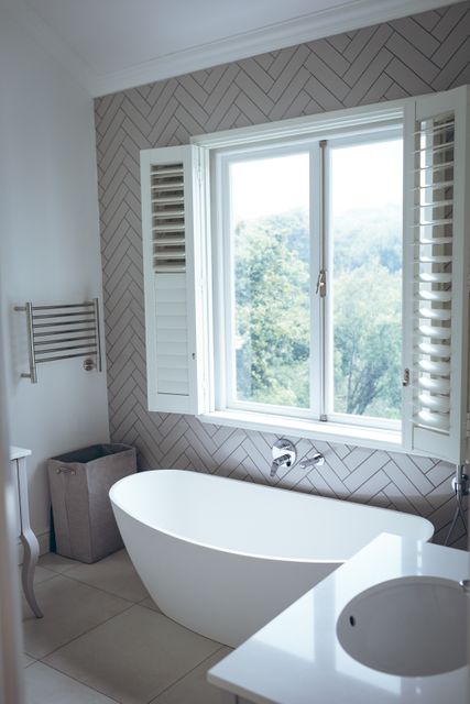 This image showcases a modern bathroom featuring a sleek freestanding bathtub positioned near a large window with shutters, offering a scenic view of lush greenery. The minimalist decor and natural light create a serene and relaxing atmosphere, making it ideal for use in home decor magazines, interior design blogs, and advertisements for luxury home products.
