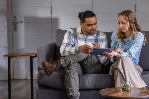 Young adults collaborating on tablet while sitting on grey sofa in cozy living room