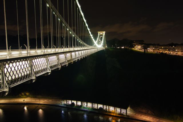 Suspension bridge glowing with lights over the river at night, showcasing architectural beauty and engineering marvel. Reflections on the water create a serene urban atmosphere. Ideal for use in travel guides, engineering concepts, urban photography projects, and promotional materials for city landmarks.