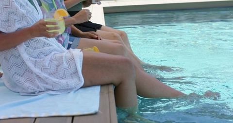 Women dipping legs in pool holding citrus cocktails on sunlit wooden deck