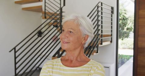 Senior woman standing with arms crossed looking upward in modern home wearing yellow striped top