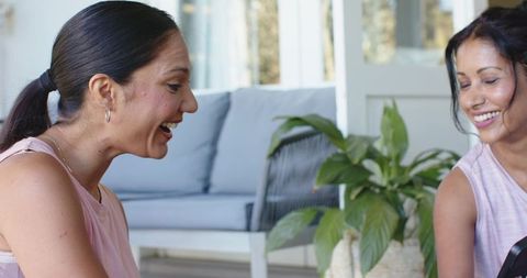 Diverse women laughing together on pale blue sofa sharing smartphone in bright living room