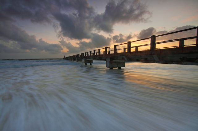 This serene scene of a pier extending over the ocean at sunset with dramatic clouds provides a tranquil coastal view. Ideal for backgrounds, wallpapers, travel brochures, posters, or social media posts, it captures the peaceful essence of a beach at twilight. The motion of the waves creates a dynamic element, making it suitable for themes related to relaxation, nature, and travel.