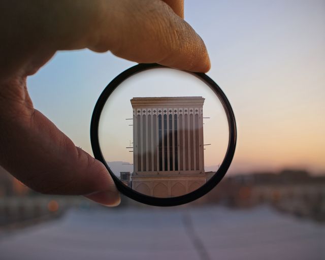 Close-up of hand holding a magnifying glass highlighting structure details of a building at dusk. Useful for architectural presentations, educational materials about structural design, and creative photography concepts displaying focus and clarity.