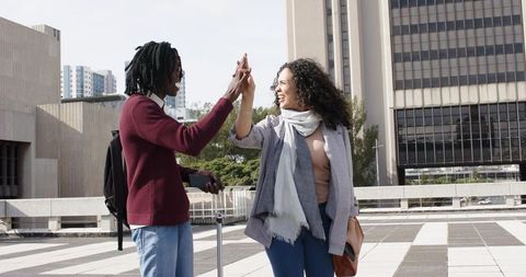 Young travelers high-fiving on urban rooftop plaza with rolling suitcase and smartphone