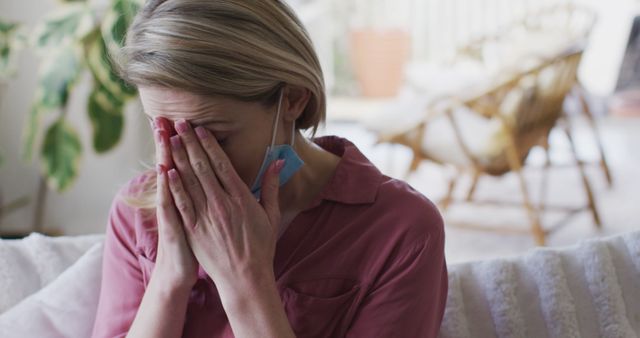 This image features a woman in a pink blouse with a face mask hanging off one ear, covering her face with her hands in apparent stress or worry. It is an evocative representation of emotional struggle during the pandemic. Ideal for articles about mental health, Covid-19 impact, stress management, and emotional well-being.