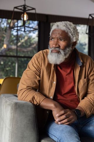 Thoughtful senior african american man sitting on sofa, leaning, holding hands near window