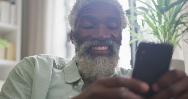 Senior man with white beard using smartphone with joy in a modern home interior. Suitable for concepts related to technology use among seniors, digital literacy, relaxation, and staying connected with family and friends. Perfect for advertising smartphone apps, technology products, or services aimed at older demographics.
