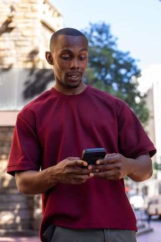 African american man standing on urban sidewalk using smartphone in maroon t-shirt