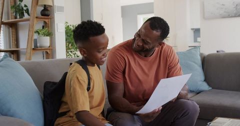 Black father and son smiling while reviewing school paper on modern living room sofa