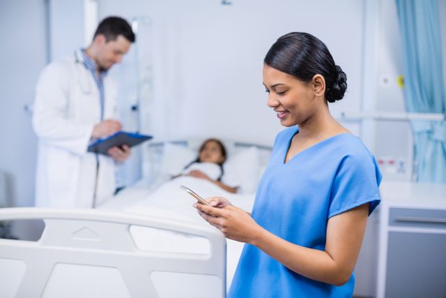 Nurse in blue scrubs smiling while using a mobile phone in a hospital room. In the background, a doctor is writing on a clipboard and a patient is lying in bed. This image can be used for healthcare, medical technology, patient care, and hospital-related content.