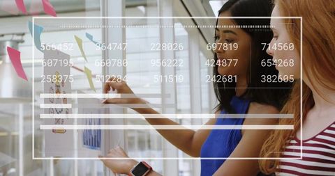 Two women collaborating at glass wall with sticky notes and analytics dashboard overlay