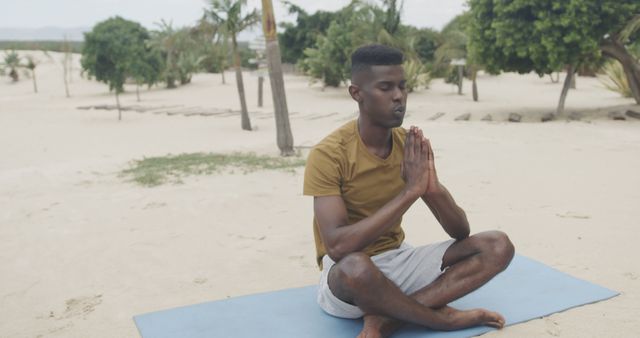 Depicts an African man sitting in a meditative pose on a sandy beach with a yoga mat. Ideal for use in wellness, meditation, and relaxation themed content. Perfect for promoting mental health, outdoor activities, yoga practices, and achieving calmness and inner peace.