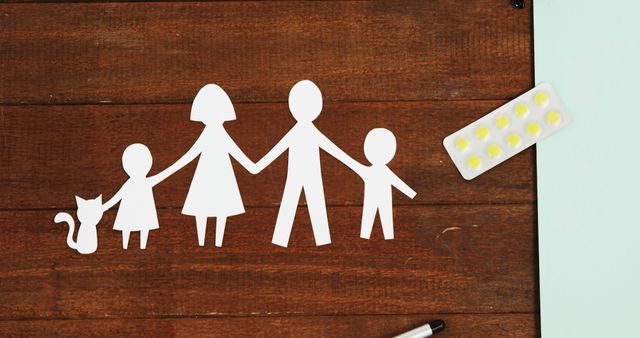 Paper cutout figures of a family including a cat are holding hands on a wooden table. Near the family are a pack of yellow pills and a pen, suggesting themes of healthcare, family planning, or medical discussions. This image can be used in parenting articles, healthcare blogs, or any content relating to family support and wellbeing.