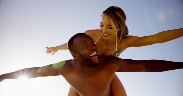 Interracial couple enjoying a sunny day at the beach. The man gives the woman a piggyback ride as they smile and have fun. Perfect for promoting summer vacations, travel destinations, joyful moments, and lifestyle blogs.