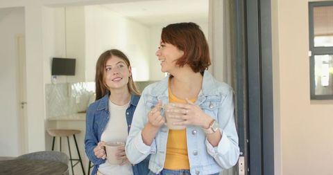 Two women enjoying morning coffee in sunlit kitchen wearing denim jackets