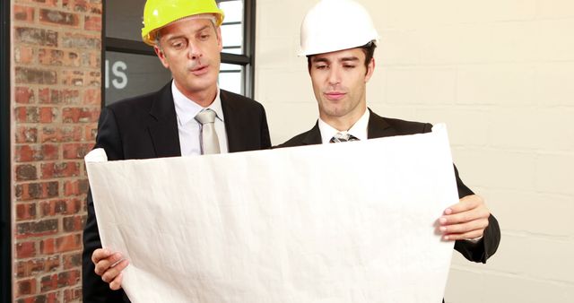 Two middle-aged Caucasian men, one in a suit and the other wearing a hard hat, are examining a blueprint together, with copy space. Their focused expressions and professional attire suggest they are involved in a construction or engineering project.