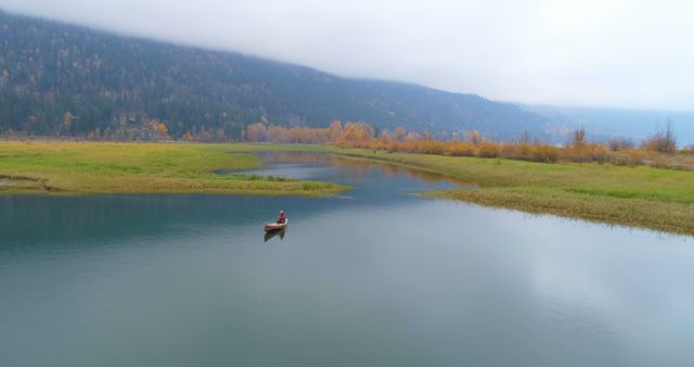 A lone kayaker explores the calm waters of a serene lake surrounded by autumn foliage and mist-covered mountains. Ideal for use in nature and outdoor activity advertisements, relaxation and wellness promotions, travel brochures, and websites focused on adventure or solo travel experiences.