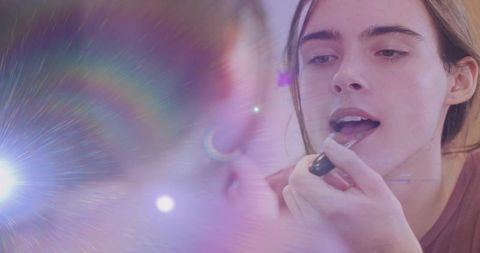 Young woman applying black lipstick in mirror with pastel lens flare and hoop earring
