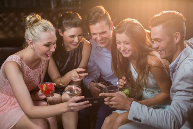 Smiling friends looking at mobile phone while having cocktail in bar