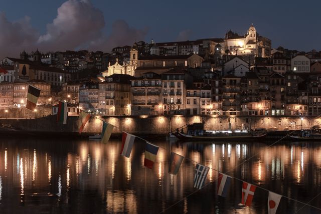 Depicts a serene night view of a picturesque European town along a river, with lights from the buildings reflecting on the water. The scene includes colorful flags hanging in the foreground and a hillside of historic, illuminated buildings. Ideal for travel magazines, tourism websites, European destination promotions, cityscape albums, and cultural heritage presentations.