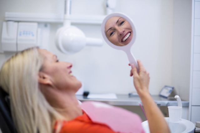 Woman sitting in dental chair, holding mirror, smiling at her reflection. Ideal for use in dental care promotions, healthcare advertisements, patient satisfaction campaigns, and oral health awareness materials.