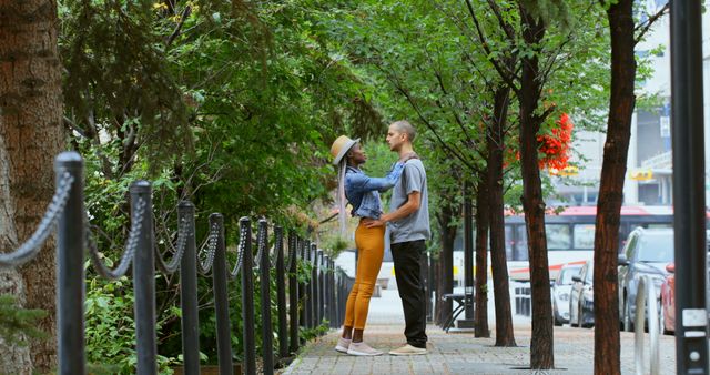The image shows a couple standing on a tree-lined sidewalk in an urban area, embracing and looking into each other's eyes, conveying a moment of tenderness and intimacy. Ideal for use in campaigns or projects centered around romance, relationships, urban living, interracial love, or outdoor summer activities. Suitable for advertisements, blog posts, or social media promotions addressing themes of romance, city life, and nature.