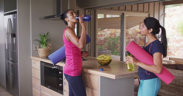 Two women at kitchen counter taking a break from yoga home session, holding water bottles and yoga mats. Ideal for content on friendship, active lifestyle, fitness routines, healthy living, and home exercise setups.