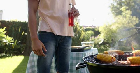 Backyard host grilling corn and burgers holding glass bottle on sunny lawn